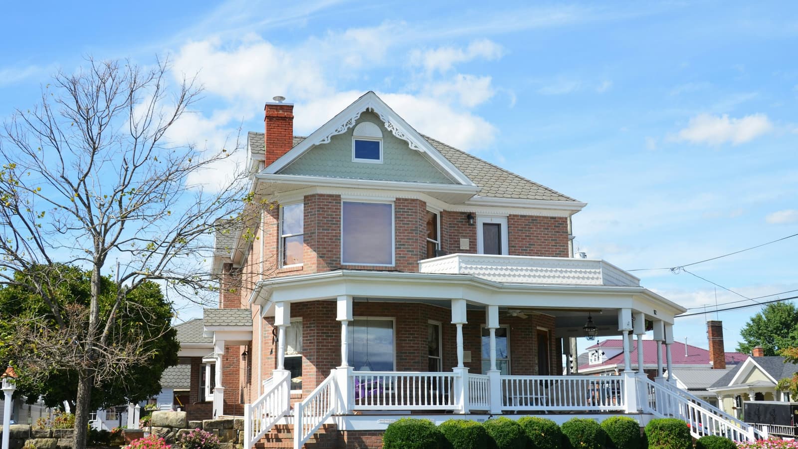 Two-story brick home with a gray asphalt-shingle roof, white wraparound porch and green lawn under a blue sky
